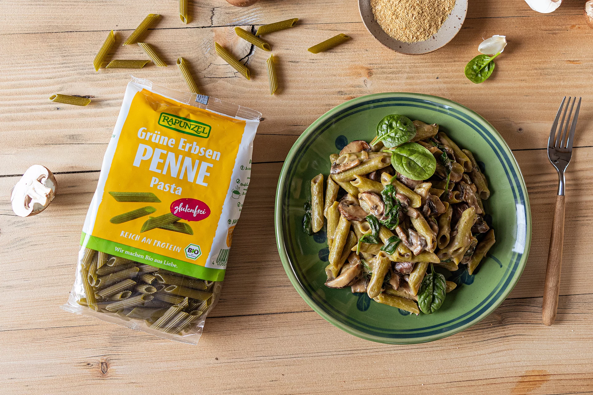 Grüne Erbsen Penne mit cremiger Pilzsauce in einer Schale, daneben offene Packung Grüne Erbsen Penne mit cremiger Pilzsauce in einer Schale serviert. Daneben liegt eine offene Packung der Grünen Erbsen Penne.