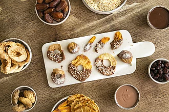 Schokolatierte Früchte mit Rapunzel Kuvertrüre gemacht Rapunel Kuvertüre halbseitig auf Rapunzel Trockenfrüchten liegend auf einem hellen Brett. Rundherum stehen kleine Schüsseln mit Trockenfrüchten und flüssiger Schokolade