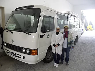 Ein durch die HAND IN HAND-Prämie finanzierter Werksbus bei CCF. Samia Chibani steht mit einer Mitarbeiterin vor dem Werksbus auf dem ein HAND IN HAND-Logo abgebildet ist.