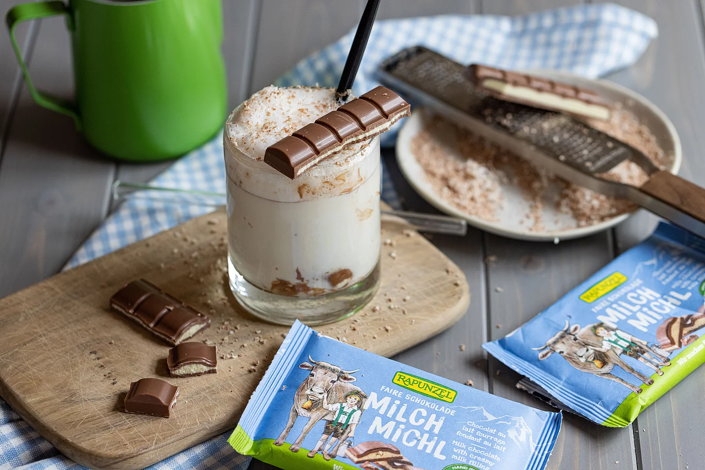 Rapunzel Milch-Michl Schokolade mit erfrischendem Getränk im Hintergrund. Eine Tafel Rapunzel Milch-Michl Schokolade liegt im Vordergrund, dahinter steht ein Glas mit einem Getränk, leicht unscharf im Hintergrund