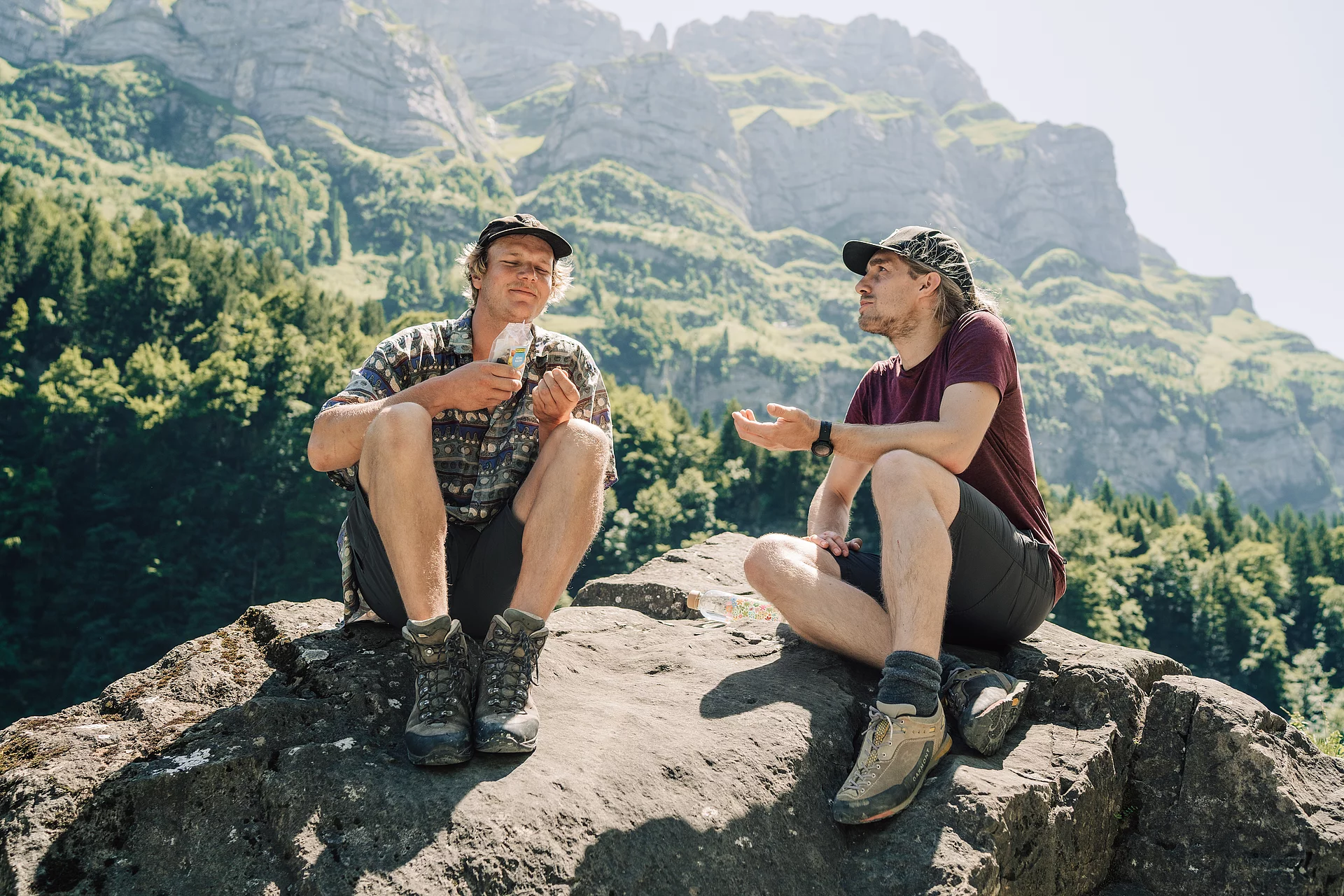Zwei Wanderer machen Pause und genießen salzige Snacks von Rapunzel. Zwei Wanderer haben sich zur Rast auf einem Großen Stein gesetzt, einer von ihnen hält eine Tüte salzige Snacks von Rapunzel in den Händen.