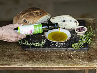 Polyphenolia Olivenöl – reich an Polyphenolen, intensiv-fruchtig, nativ extra Rapunzel Bio-Olivenöl Polyphenolia wird in ein Schälchen gegossen, im Hintergrund Olivenbrot und Holzbrettchen – reich an Polyphenolen, kräftig-fruchtiger Geschmack