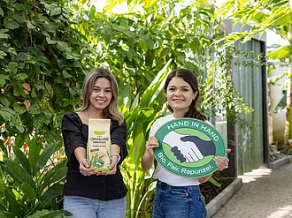 HAND IN HAND-Partner Manduvira mit Zuckerrohr. Die Leiterinnen des HAND IN HAND-Partners Manduvira mit Zuckerrohr, einer Packung Rohrzucker und dem HAND IN HAND-Logo.