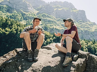 Zwei Wanderer machen Pause und genießen salzige Snacks von Rapunzel. Zwei Wanderer haben sich zur Rast auf einem Großen Stein gesetzt, einer von ihnen hält eine Tüte salzige Snacks von Rapunzel in den Händen.
