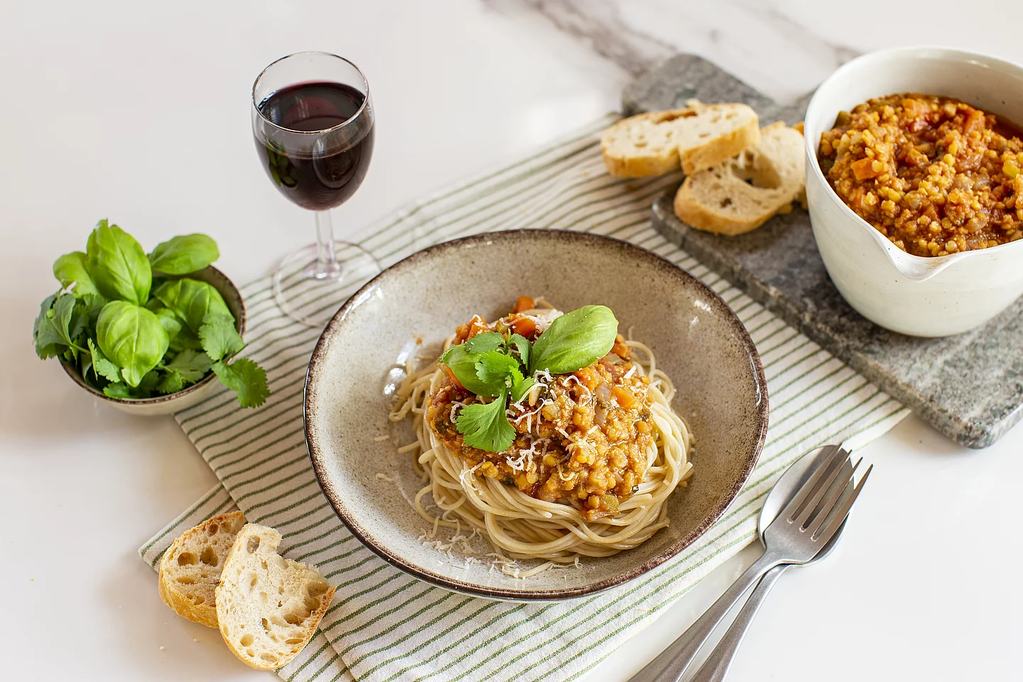 Vegane Linsen-Bolognese mit Spaghetti und frischen Kräutern Ein tiefer Keramikteller mit Spaghetti und einer herzhaften Linsen-Bolognese, garniert mit frischem Basilikum und Koriander. Im Hintergrund stehen eine Schüssel mit der restlichen Sauce aufgeschnittenes Baguette, ein Glas Rotwein und eine kleine Schale mit Kräutern auf einer hellen Tischoberfläche