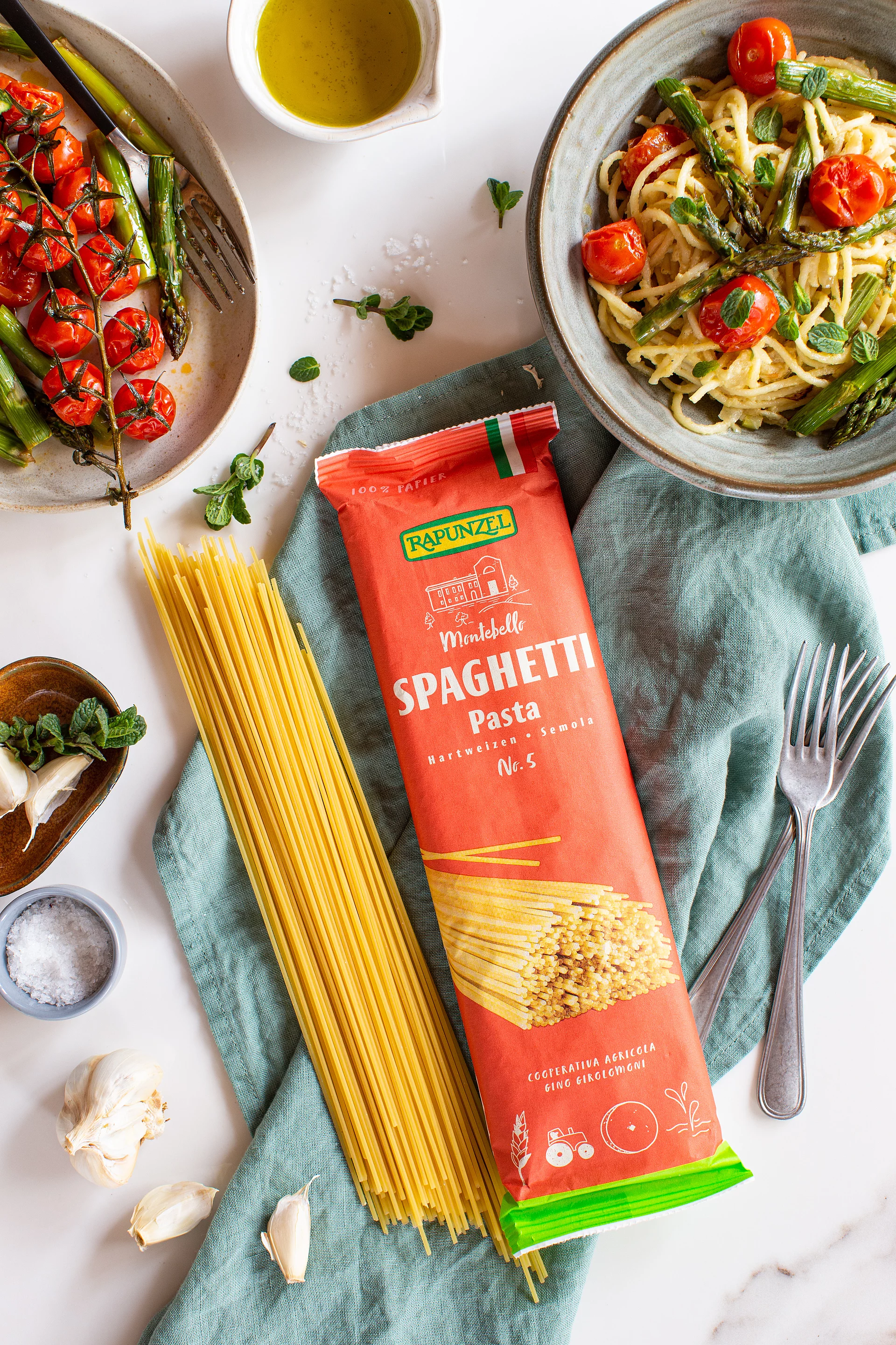 Rapunzel Bio-Spaghetti in Originalverpackung und lose auf Küchentuch, angerichtetes Nudelgericht mit Gemüse drumherum. Rapunzel Bio-Spaghetti werden sowohl in der Originalverpackung als auch lose auf einem Küchentuch präsentiert. Drumherum ist ein angerichtetes Nudelgericht mit Gemüse dekorativ angerichtet.
