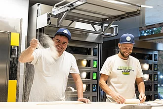 Mehle und Spezialmehle nutzt man auch in der Rapunzel Bäckerei. Zwei Bäcker am Backtisch in der Rapunzel Bäckerei bei der Arbeit, einer wirft Mehl in die Luft, beide lachen.