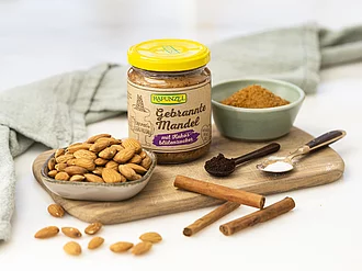 Retromus Gebrannte Mandel – Einfachheit in ihrer reinsten Form Glas Rapunzel Retromus Gebrannte Mandel auf Küchenoberfläche, daneben die Zutaten wie Mandeln und Rohrzucker – wenige, natürliche Bio-Zutaten für authentischen Genuss