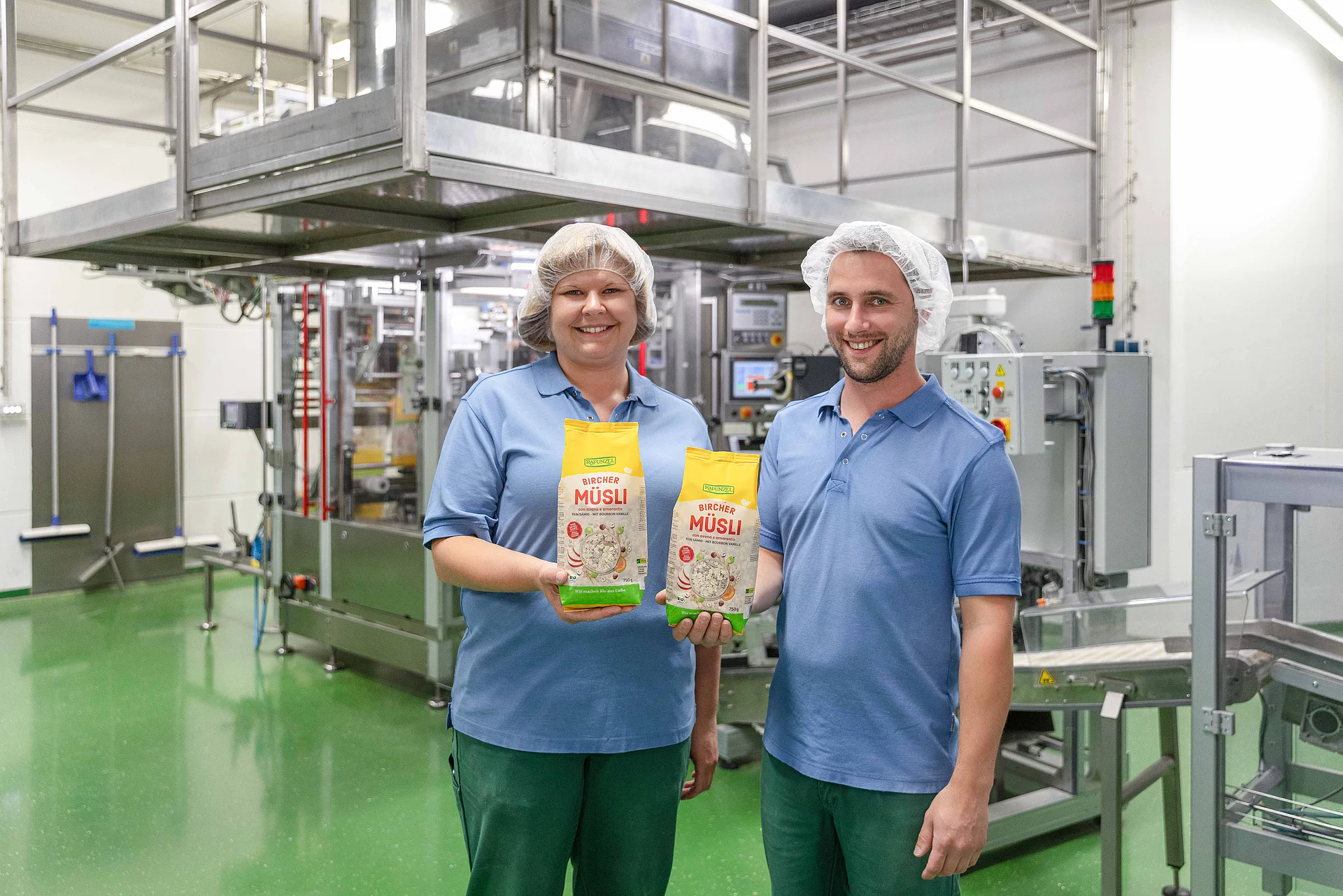 Rapunzel Produktionsmitarbeiter stellen Müsli her. Zwei Mitarbeiter aus der Rapunzel Produktion halten je eine Packung Müsli in die Kamera, im Hintergrund die Müslianlage.