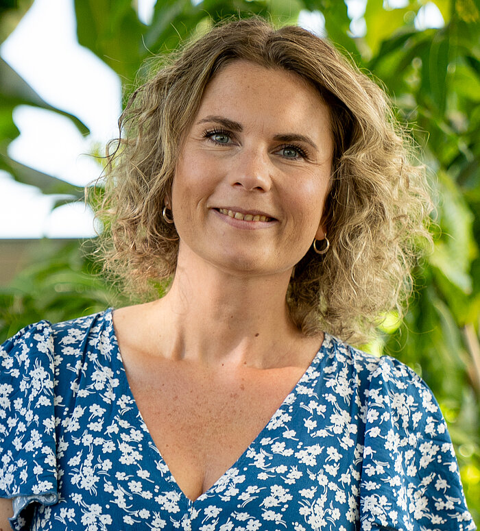 Anne Schubert Porträt von Anne Schubert aus dem Rapunzel Kundenservice, lächelnd in die Kamera vor tropischen Pflanzen