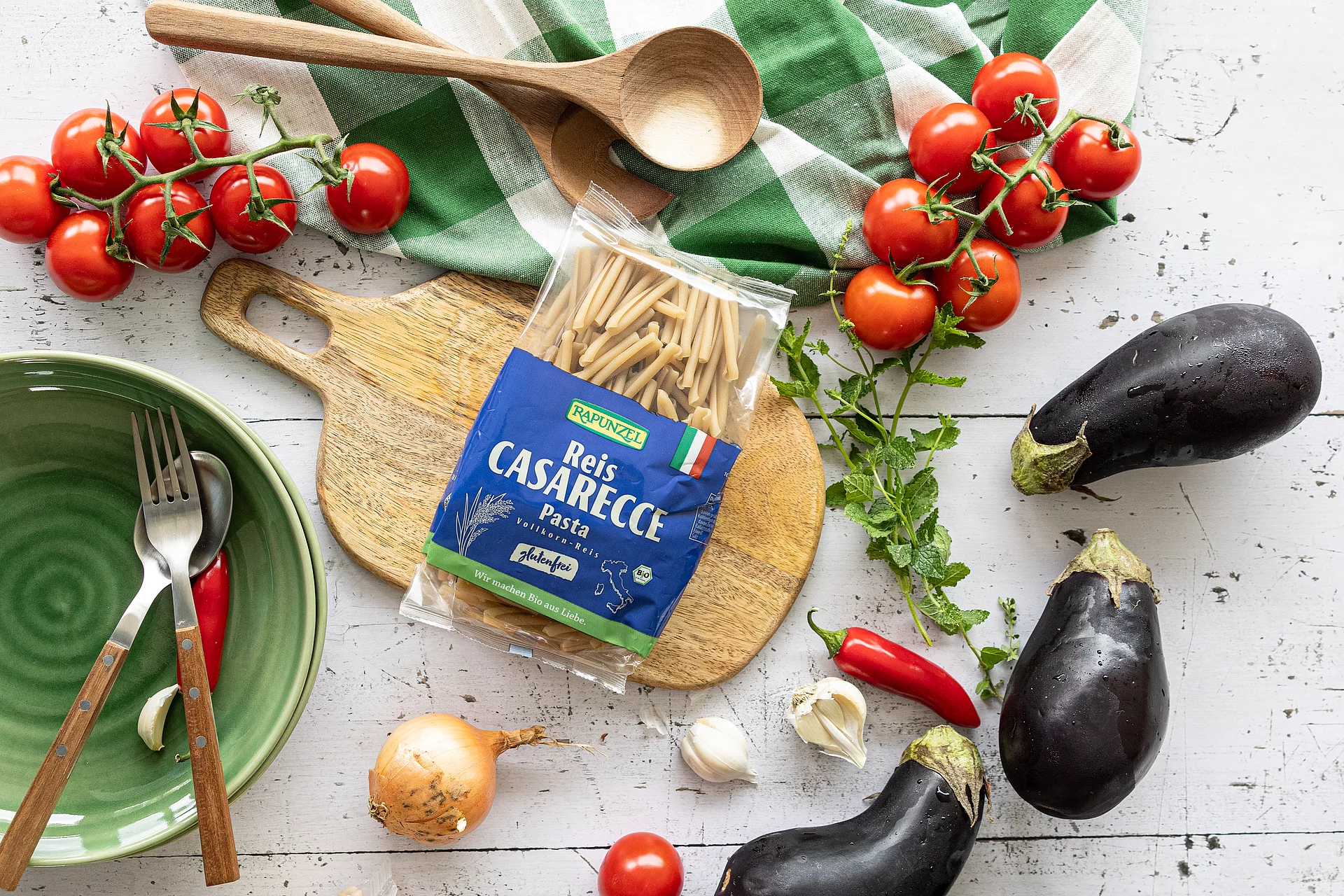 Reis Casarecce Pasta glutenfrei auf Schneidebrett mit Gemüse und Küchenutensilien. Packung Reis Casarecce Pasta glutenfrei auf einem Schneidebrett, umgeben von Tomaten, Aubergine, Chilischoten, Knoblauch, Minze, Zwiebel, einem grün-weißen Küchentuch, Holzlöffel sowie einer grünen Schüssel mit Gabel und Löffel.