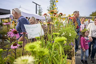 Eine Gruppe von Menschen, jung wie alt, die sich neben blühenden Pflanzen aufhalten. Es herrscht eine zufriedene und friedliche Stimmung.