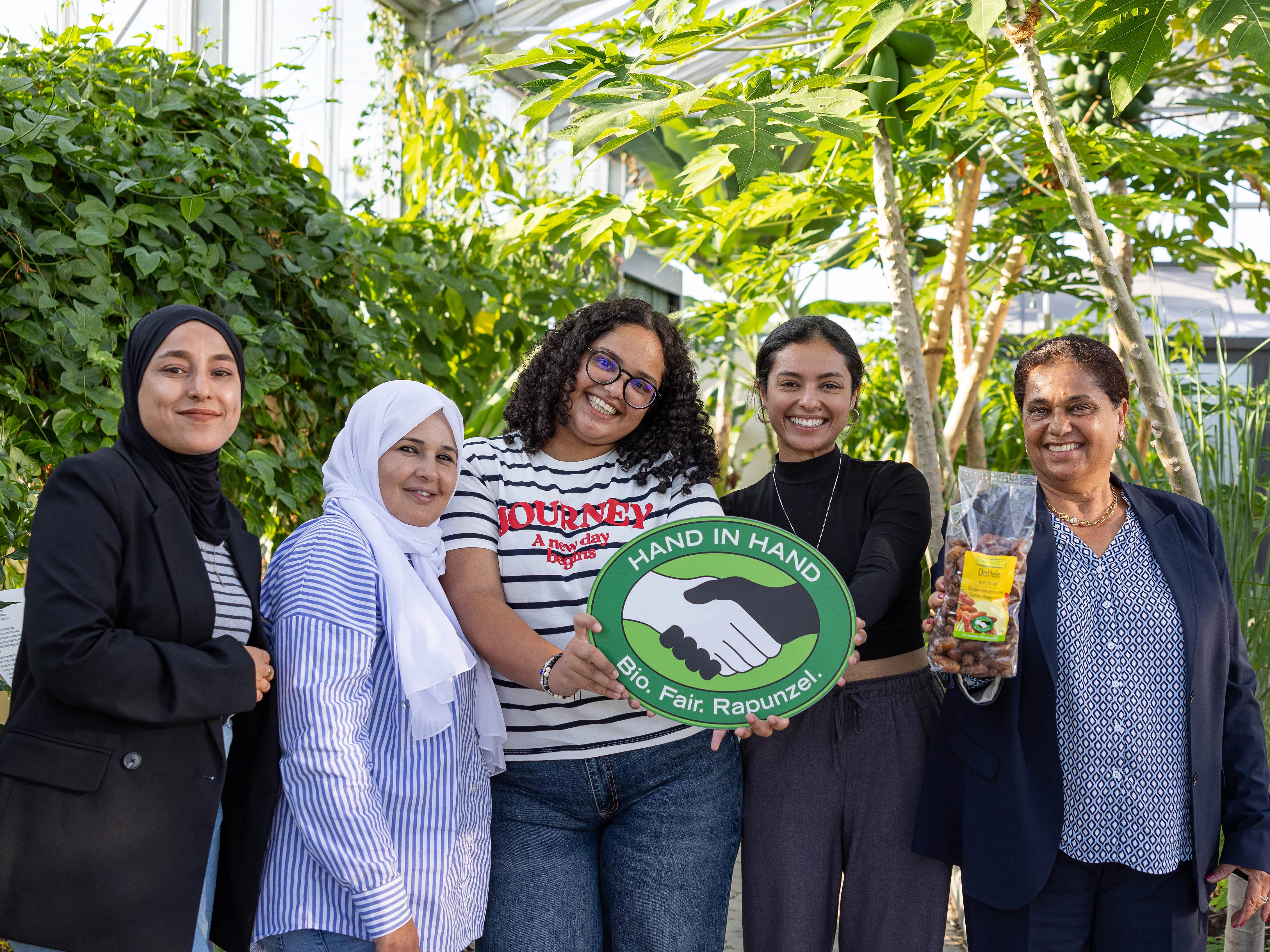 CCF zu Besuch bei Rapunzel in Legau. Gruppenfoto im Tropenhaus von Rapunzel mit CCF-Gründerin Samia Chibani. Mit im Bild sind das HAND IN HAND-Logo und die Datteln, die von Rapunzel verkauft werden.