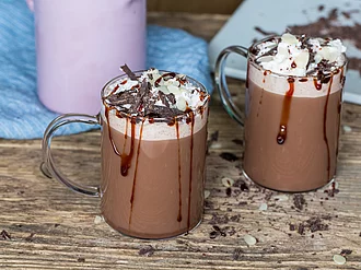 Zwei tassen Heisse Schokolade mit Sahne Auf einem Holztisch stehen zwei Tassen heiße Schokolade mit Sahne und Schokoladensauce garniert.Im Hintergrund steht, leicht angeschnitten, Küchengeschirr