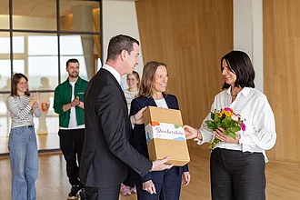 Rapunzel Geschenkpaket für Mitarbeiter oder Kunden. Drei Menschen stehen im Vordergrund. Es wird ein Rapunzel Dankespaket überreicht. Im Hintergrund zwei klatschende Menschen.